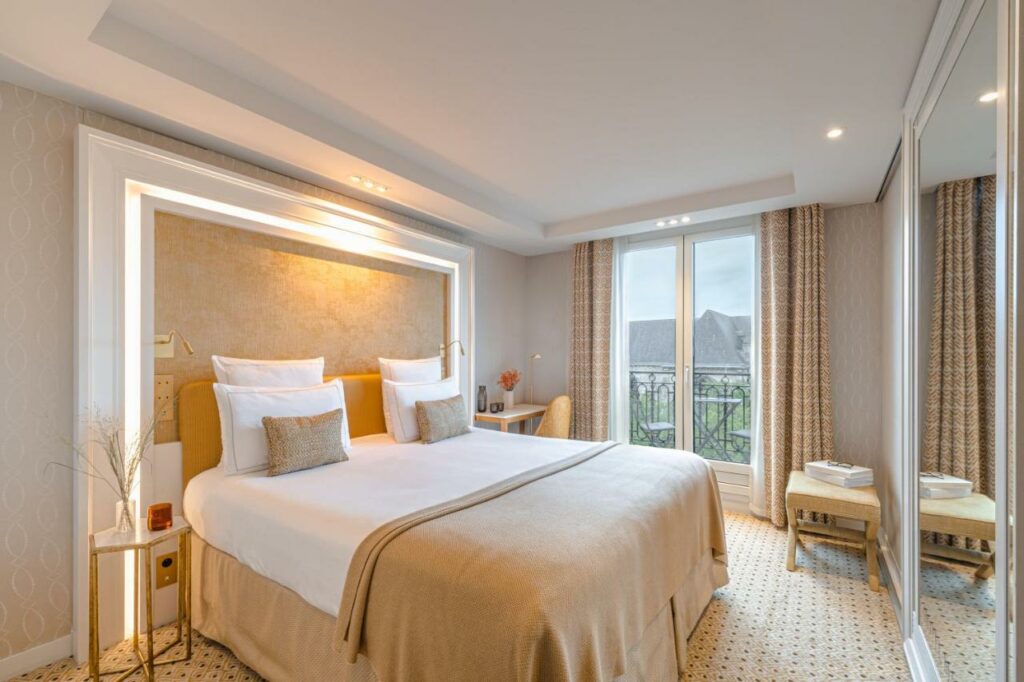 Quarto do Hôtel Madison, um dos hotéis em Saint Germain em Paris. Uma cama de casal está encostada na parede esquerda do local com mesinhas de cabeceira e abajures dos dois lados. Logo em frente fica um guarda-roupa com espelho. Na lateral direita há uma grande janela com cortinas na parede, e uma mesa de trabalho com cadeira, vaso de flores e garrafa d'água fica ao lado.