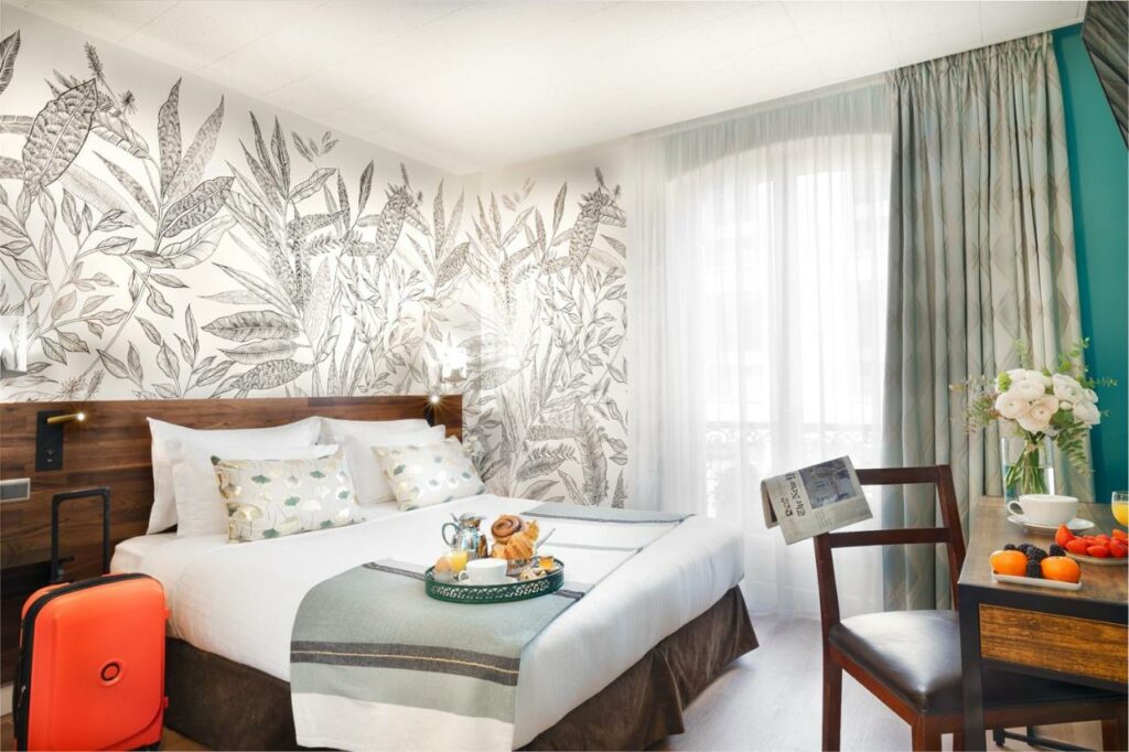 Quarto do Hôtel Delavigne, um dos hotéis em Saint Germain em Paris. Uma cama de casal está encostada na parede esquerda do local com mesinhas de cabeceira e abajures dos dois lados, assim como uma bandeja com itens de café da manhã aos pés. Logo em frente fica uma mesa com cadeira, jornal, vaso de flores e outros alimentos do café. Na lateral direita da cama há uma janela com cortinas.
