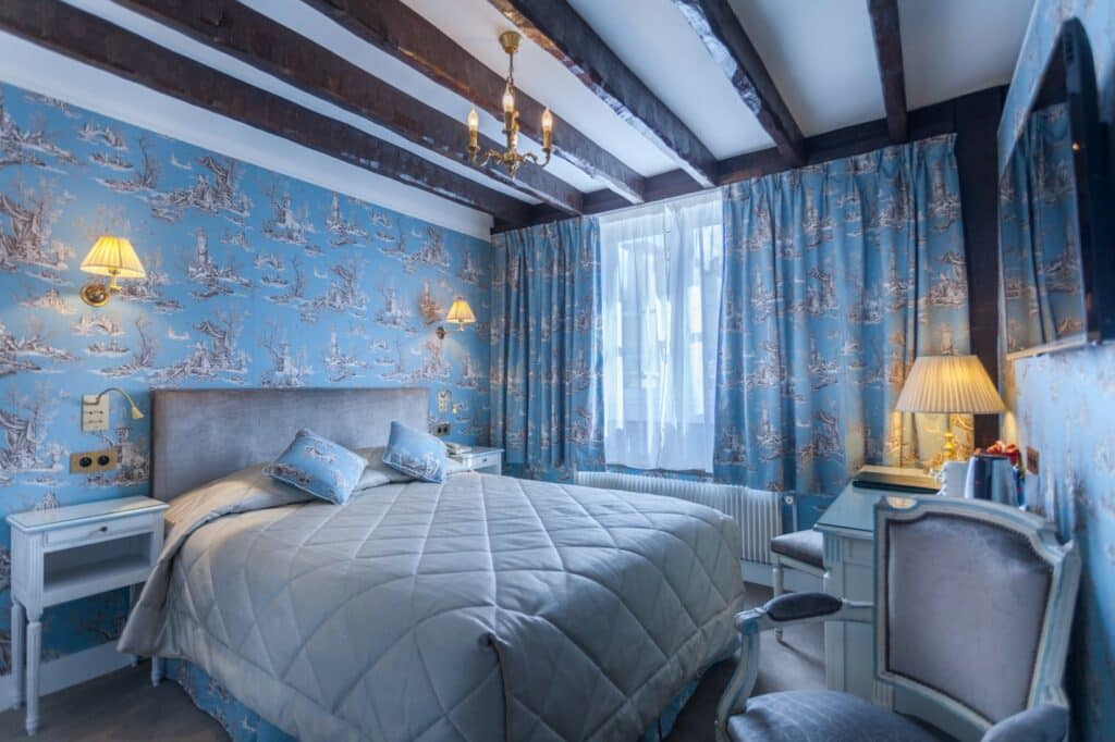 Quarto do Grand Hôtel de L'Univers Saint-Germain, um dos hotéis em Saint Germain em Paris. Uma cama de casal está encostada na parede esquerda do local com mesinhas de cabeceira e abajures dos dois lados. Logo em frente fica uma mesa com banquinho, abajur e chaleira, e ao lado há uma cadeira. Na lateral direita há uma janela com cortinas.