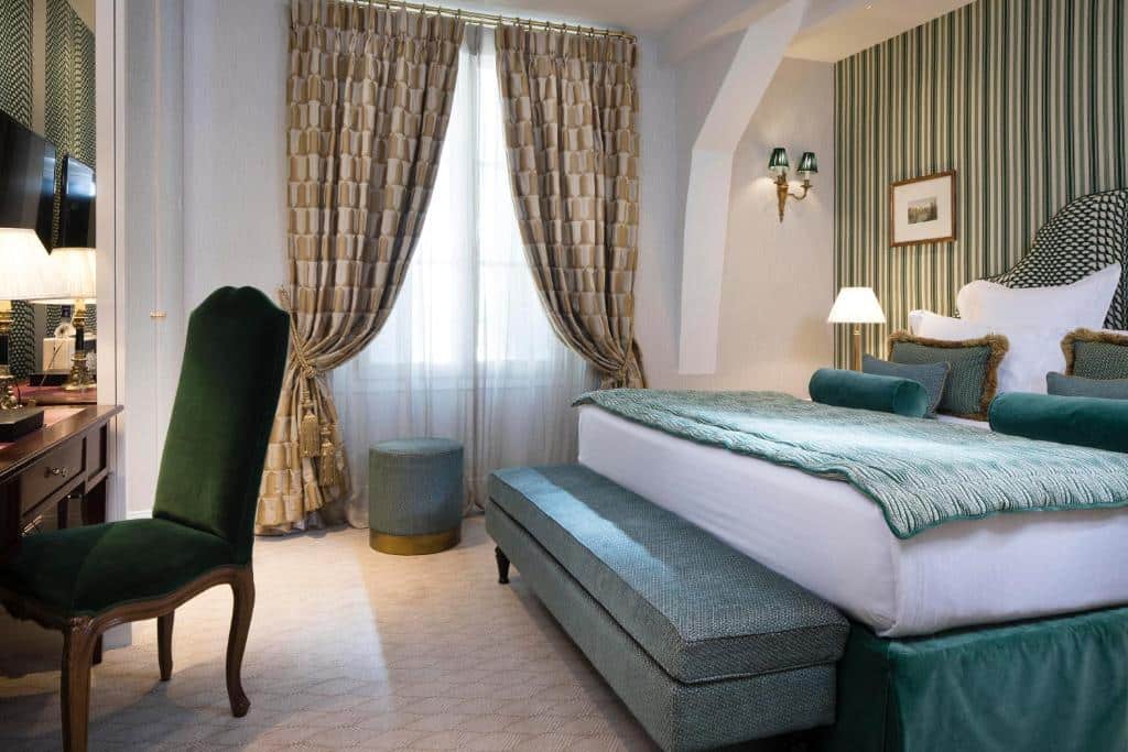 Quarto do Relais Christine, um dos hotéis em Saint Germain em Paris. Uma cama de casal repleta de almofadas e com um banco aos pés fica na lateral direita da foto, e há mesinhas de cabeceira com abajur dos dois lados. Em frente à ela fica uma mesa de trabalho com cadeira e TV.Ao lado esquerdo da cama há uma janela com cortinas e um banquinho em frente.