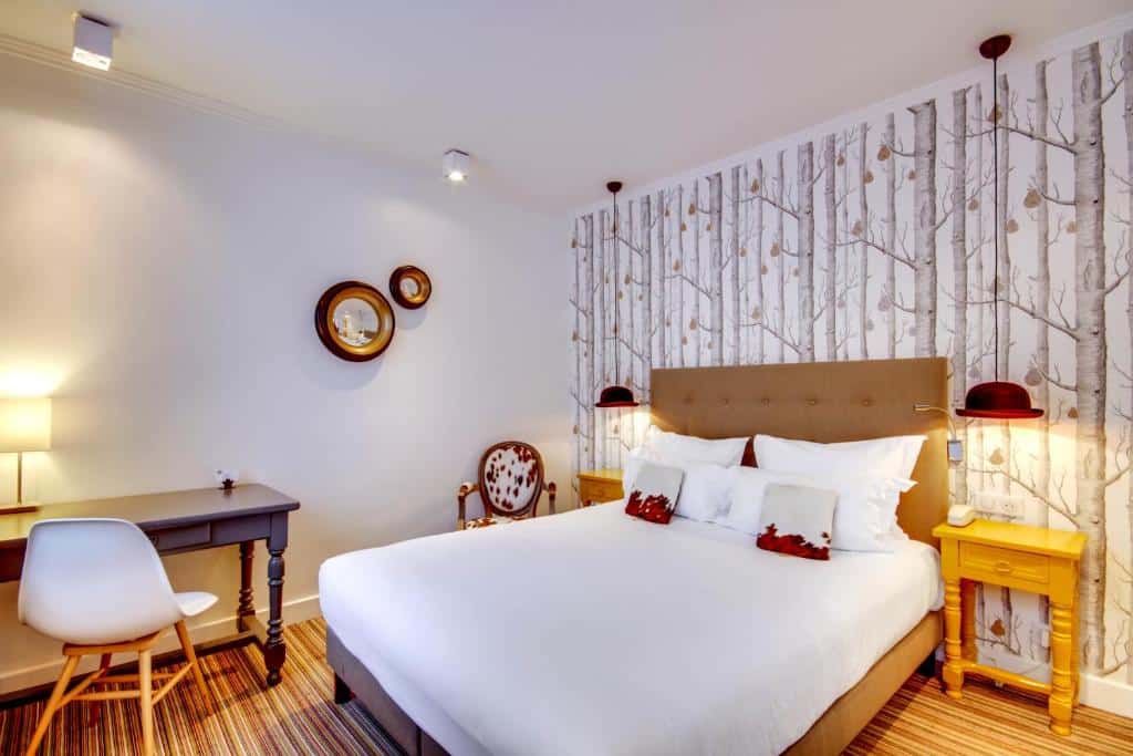 Quarto do Hôtel Sèvres Saint Germain, um dos hotéis em Saint Germain em Paris. Uma cama de casal está encostada na parede direita do local com mesinhas de cabeceira e abajures dos dois lados, assim como uma poltrona encostada na parede lateral. Em frente à poltrona fica uma mesa com cadeira e abajur.