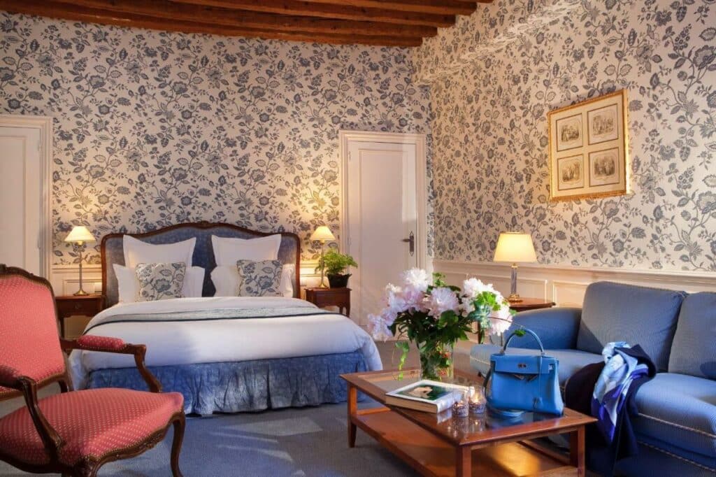 Quarto do Hotel d'Angleterre, um dos hotéis em Saint Germain em Paris. Uma cama de casal está encostada na parede central com mesinhas de cabeceira e abajures dos dois lados. Logo em frente fica uma área de estar com mesa, sofá e poltrona. Na lateral direita há outra mesinha com abajur e um quadro acima.