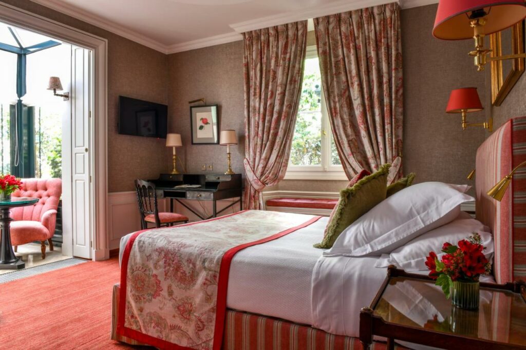 Quarto do Hôtel de l'Abbaye, um dos hotéis em Saint Germain em Paris. Uma cama de casal está encostada na parede direita do local com mesinhas de cabeceira e abajures dos dois lados. Logo em frente fica um terraço com área de estar com mesa e duas poltronas. Na lateral direita da cama há uma janela com cortinas na parede, e uma mesa de trabalho com cadeira e abajures fica ao lado esquerdo da janela. Há também uma TV ao lado da mesa.