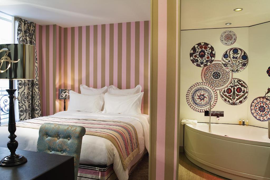 Quarto do Hôtel Le Bellechasse Saint-Germain, um dos hotéis em Saint Germain em Paris. Uma cama de casal está encostada na parede esquerda do local com mesinhas de cabeceira e abajures dos dois lados. Logo em frente fica uma mesa com cadeira e abajur.Há uma parede dividindo o quarto do banheiro, onde é possível ver uma banheira de hidromassagem.