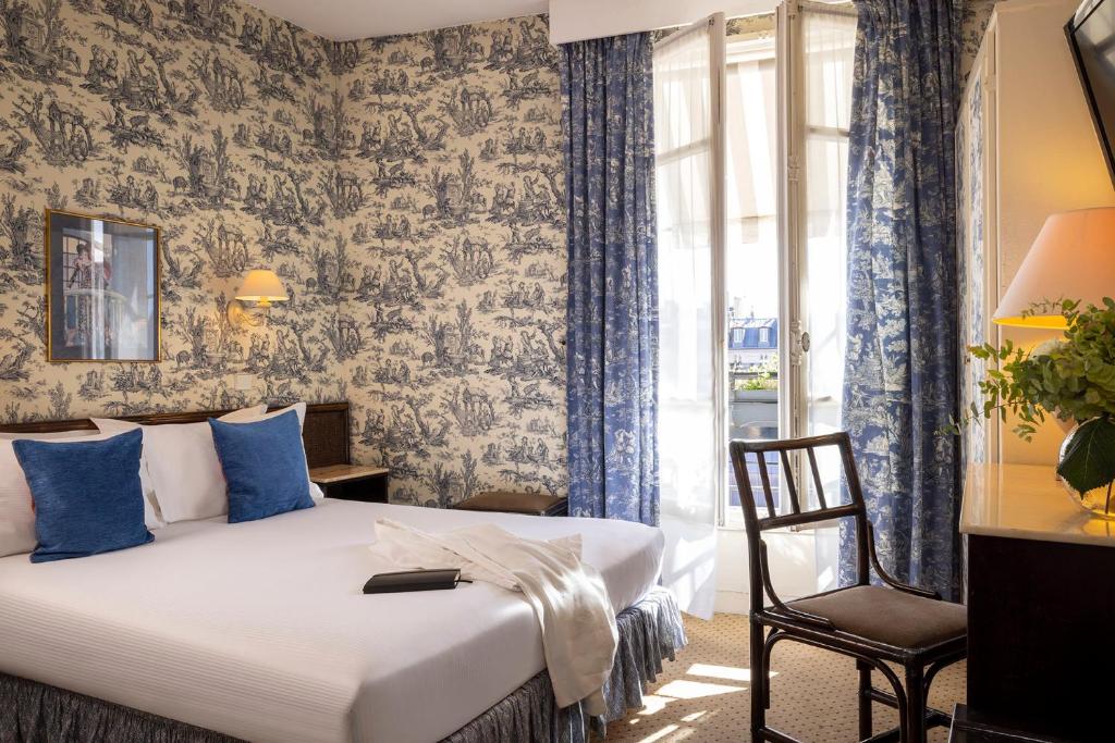 Quarto do Hotel Clément, um dos hotéis em Saint Germain em Paris. Uma cama de casal está encostada na parede esquerda do local com mesinhas de cabeceira e abajures dos dois lados, assim como um quadro na parede acima da cabeceira. Logo em frente fica uma mesa com cadeira, vaso de plantas, abajur e TV na parede acima. Na lateral direita há uma janela com cortinas.