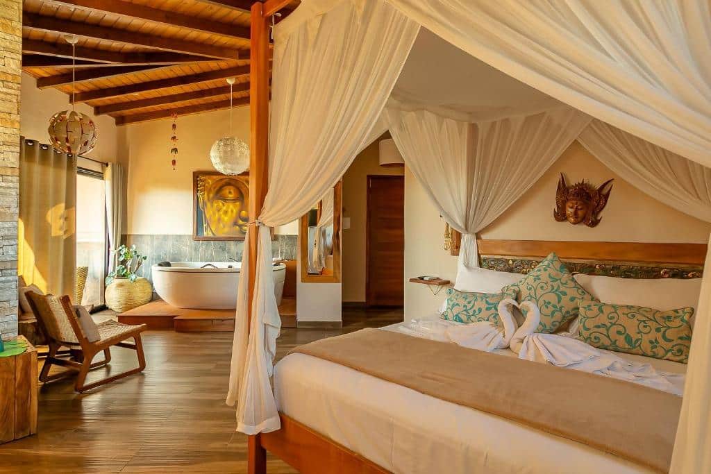 Quarto do hotel Shiva Boutique Hotel - Praia do Rosa. Uma cama de casal com dossel está do lado direito. Ao fundo, no lado esquerdo, há uma cadeira e uma banheira encostada na parede, abaixo de um quadro e ao lado de uma porta transparente com cortinas e um vaso de planta. No meio da imagem, entre a banheira e a cama, é possível ver um espelho na parede e uma porta fechada ao fundo. Esse um dos hotéis em Santa Catarina.