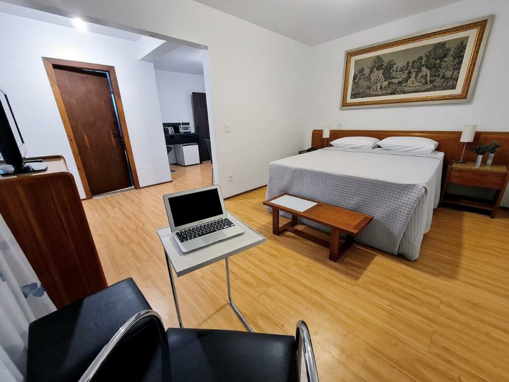 Quarto do Hotel Germânia. Uma cama de casal está no meio e um banquinho de madeira está na sua ponta. Do lado direito da cama há uma mesinha. Em frente a ela está uma mesa de apoio com um notebook, uma poltrona e uma tv. Ao fundo é possível ver uma porta. Foto para ilustrar o post de hotéis em Santa Catarina.