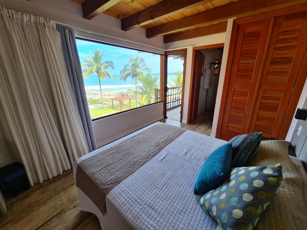 Quarto do Hotel Atalaia do Mariscal. Uma cama de casal está de frente para uma grande janela de vidro com vista para a praia. Existem também cortinas. Do lado direito estão duas portas, uma do banheiro e outra levando para a varanda. Foto para ilustrar o post de hotéis em Santa Catarina.