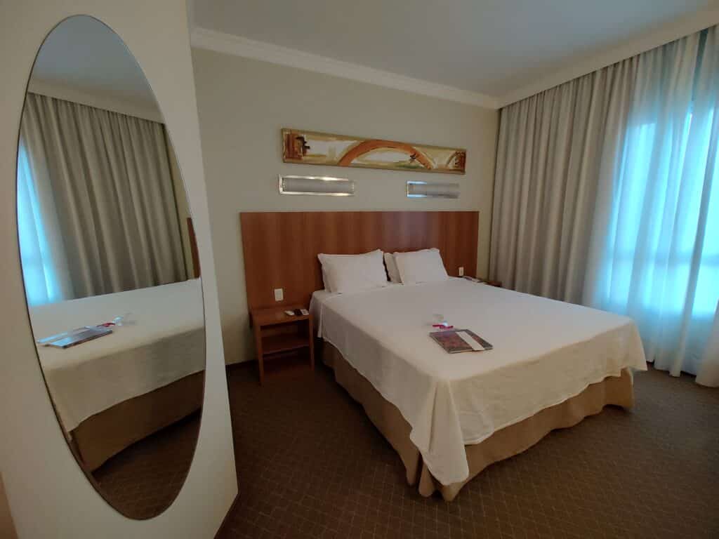 Quarto do Bourbon Joinville Convention Hotel. Uma cama de casal está no canto direito, perto da janela com cortinas. Ao lado esquerdo da cama há uma mesinha e mais a frente um espelho de forma oval. Foto para ilustrar o post de hotéis em Santa Catarina.