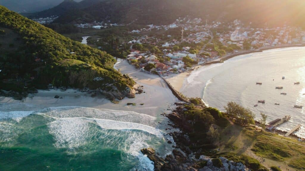 Vista de cima da Praia da Armação, em Florianópolis. É possível ver o mar, alguns barcos dentro dele e muito vegetação em volta. Ao fundo existem também várias casas.