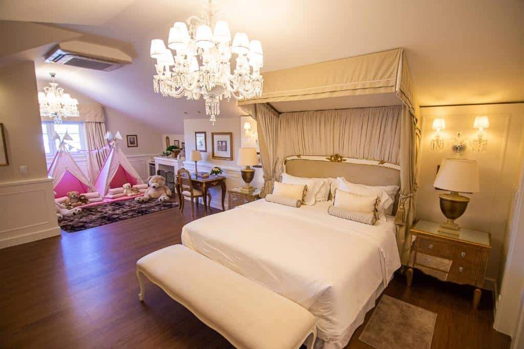 Quarto do Villa do Vale Boutique Hotel. Uma cama de casal com dossel e um recamier na ponta está no lado direito da imagem, ao lado de uma mesinha com abajur. Na parte esquerda, após uma mesa pequena com uma cadeira, há uma área infantil. Duas cabanas rosas com alguns ursos e almofadas estão em frente a janela. Esse é um dos hotéis em Santa Catarina, na cidade de Blumenau.