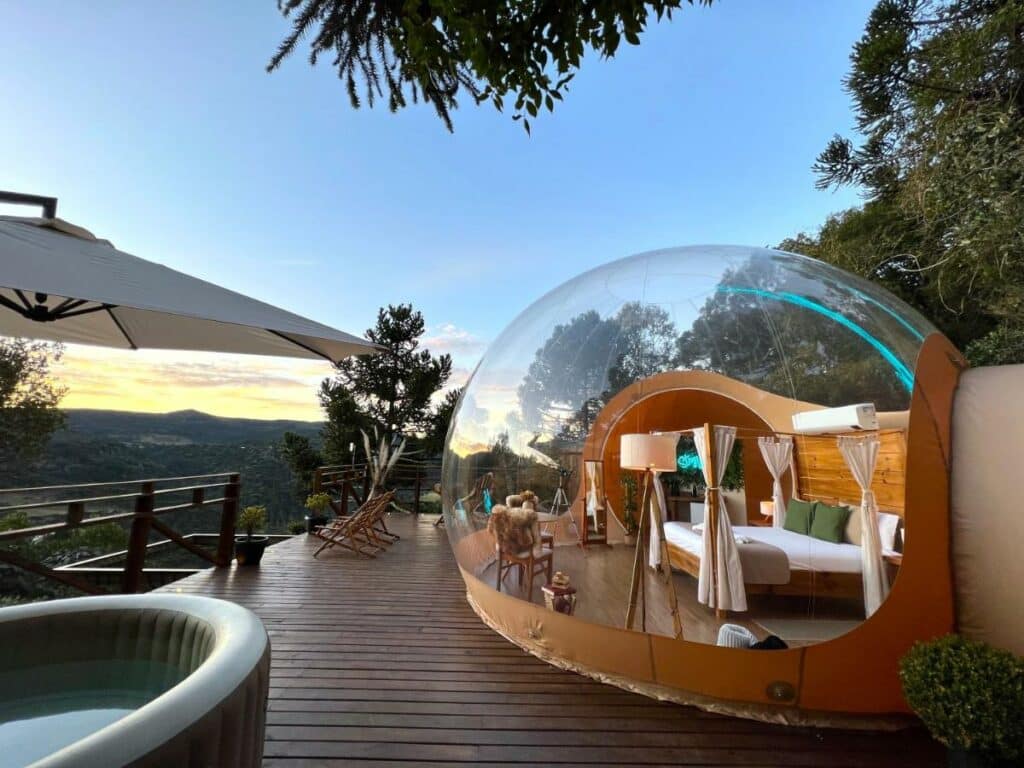 Vista geral do quarto e varanda do hotel Zion Bubble Glamping. O quarto está dentro de uma bolha transparente, que possibilita ver o céu e toda a paisagem do local. Uma cama de casal com dossel está no meio e algumas coisas como uma cadeira, espelho e um telescópio estão ao redor. Um ar-condicionado está acima da cama. Fora da bolha é possíver ver uma jacuzzi com guarda-sol, duas cadeiras de madeira e a vegetação como paisagem. Este é mais um dos hotéis em Santa Catarina.
