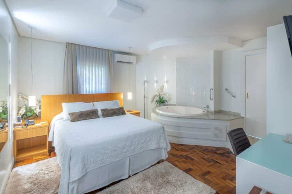 Quarto do Hotel Glória. Uma cama de casal está ao lado esquerdo, do lado de uma mesinha colada na parede. Em frente a cama há uma mesa com uma cadeira. Do lado direito há uma banheira de hidromassagem, ela está em uma área elevada, mas existem duas barras de apoio na parede. Essa é uma foto para ilustrar o post de hotéis em Santa Catarina.