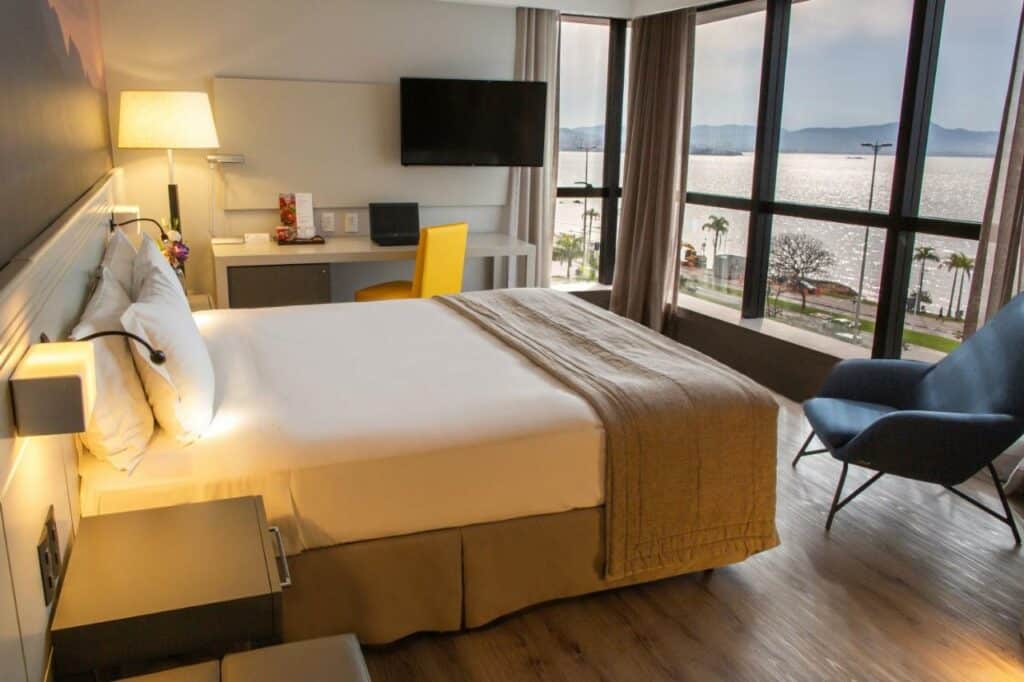 Quarto do hotel Novotel Florianopolis. Uma cama de casal está no meio, de frente para uma cadeira e uma parede transparente com vista para a rua e para o mar. Em um lado do quarto está uma mesa, uma cadeira, abajur e a televisão, pendurada na parede. Do outro lado há uma mesa com gaveta. Esse é um dos hotéis em Santa Catarina.
