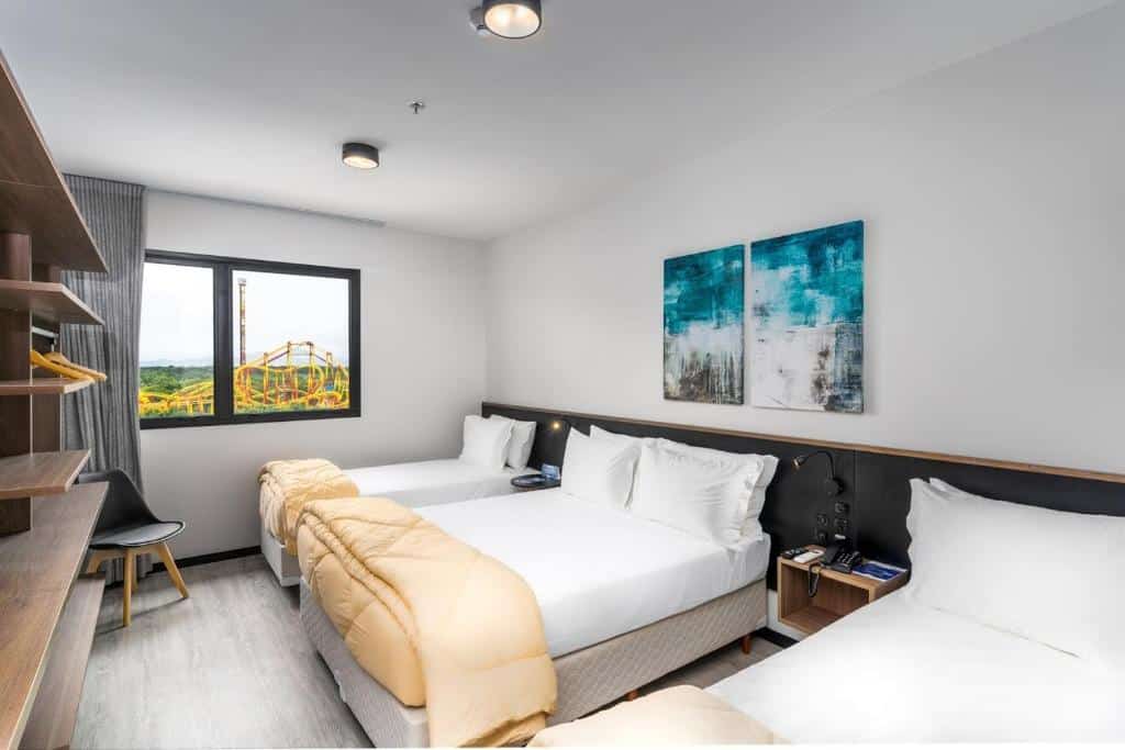 Quarto do hotel Solar Pedra da Ilha - OFICIAL. Ele possui três camas, com uma de casal no meio e duas outras em cada lado. Na parede de fundo há uma janela com vista para uma montanha-russa. Na parede de frente para as camas há um móvel com várias prateleiras. Este também é um dos hotéis em Santa Catarina, em Penha.