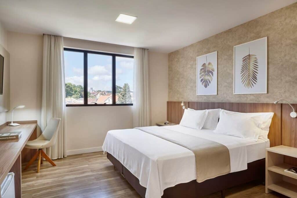 Quarto do Lefel Hotel, em São Bento do Sul. Uma cama de casal está no meio, ao lado de uma mesinha. Na parede do lado esquerdo há uma janela e cortinas. Já na parede de fente para a cama há uma mesa com uma cadeira e uma tv no alto. Foto para ilustrar o post de hotéis em Santa Catarina.
