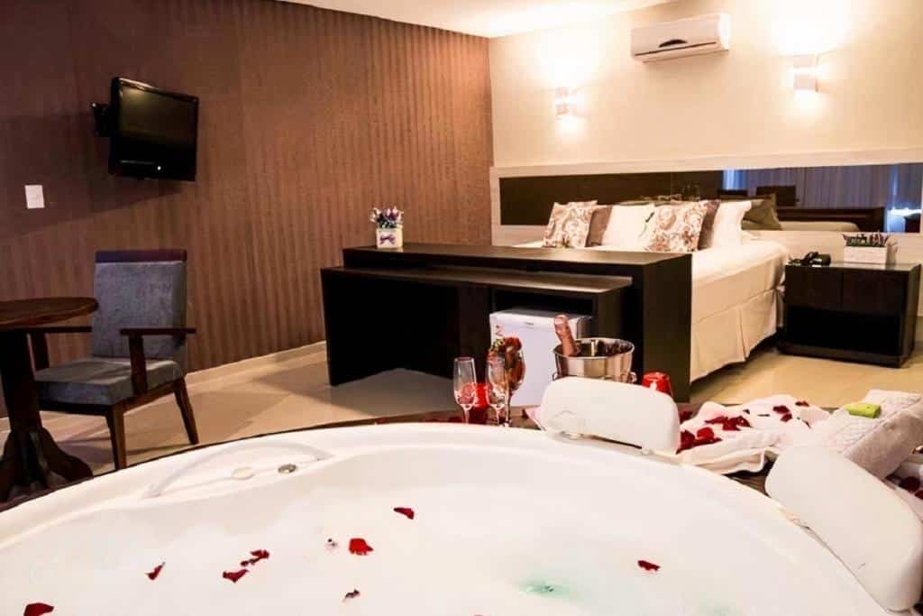 Quarto do Reserva Praia Hotel. Em primeiro plano há uma banheira de hidromassagem coberta de pétalas de rosa e com um balde de champagne e taças. Em segundo plano está uma cama de casal, uma mesinha ao seu lado e um ar-condicionado na parede. Em outra parede está uma tv. Perto da cama e da banheira há uma mesa e uma cadeira. Foto para ilustrar o post de hotéis em Santa Catarina.