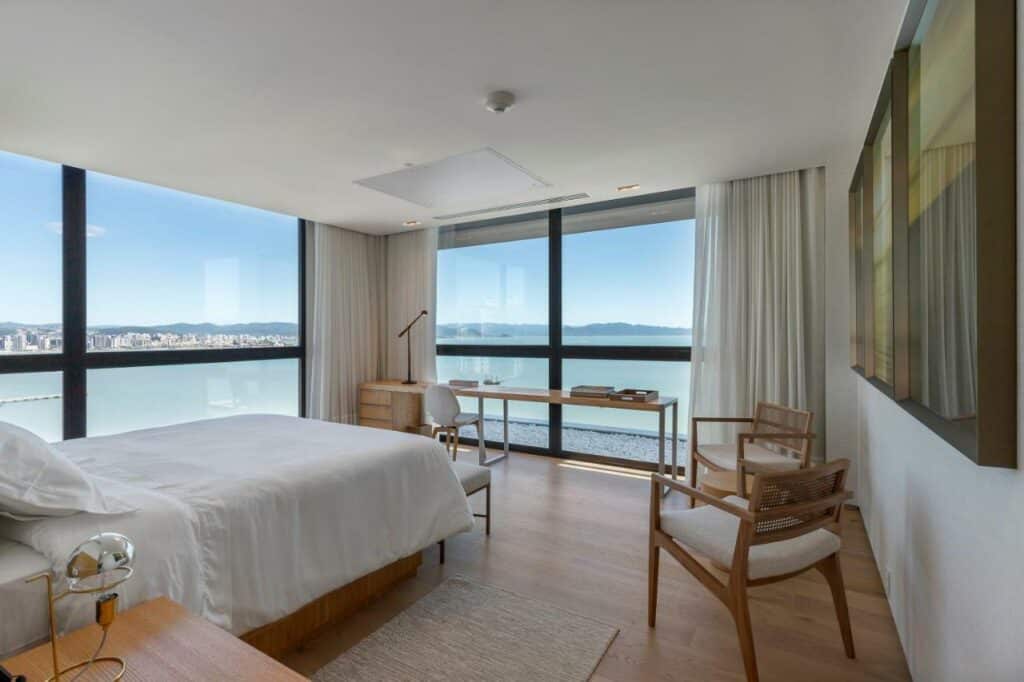Quarto do hotel LK Design Hotel Florianópolis. Uma cama de casal está no meio, ao seu lado há uma mesinha e em sua ponta um banquinho. As paredes de frente para a cama e ao seu lado esquerdo são de vidro e têm uma vista paronâmica do mar. Ao redor da cama e perto do vidro em frente a ela estão uma mesa e algumas cadeiras. Este é um dos melhores hotéis em Santa Catarina.