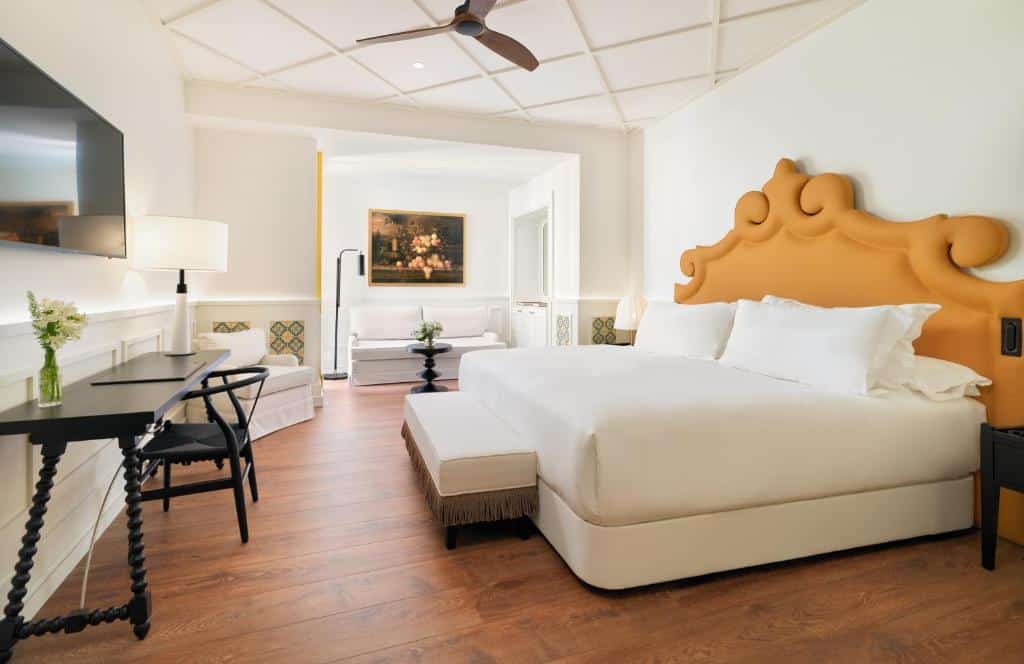 Quarto espaçoso do H10 Casa de la Plata. Uma cama de casal com uma cabeceira amarela no canto direito. De frente uma mesa de trabalho e uma televisão. No fundo um sofá e uma luminária. Foto para ilustrar post sobre Hotéis em Sevilha.