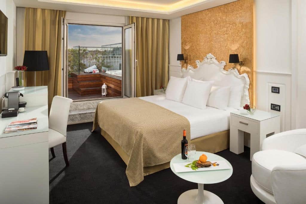 Quarto do Hotel Colón Gran Meliá - The Leading Hotels of the World. Uma mesa de centro com vinho e uma poltrona. Uma cama de casal atrás e de frente uma mesa de trabalho com televisão. Atrás a sacada com uma jacuzzi.