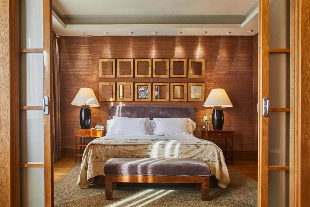 Quarto do Silken Al-Andalus Palace. Uma porta de madeira dos dois lados aberta, uma cama de casal com uma luminária de cada lado. Na frente da cama um banco estofado. Diversos quadrinhos em cima da cabeceira da cama. Foto para ilustrar post sobre Hotéis em Sevilha.