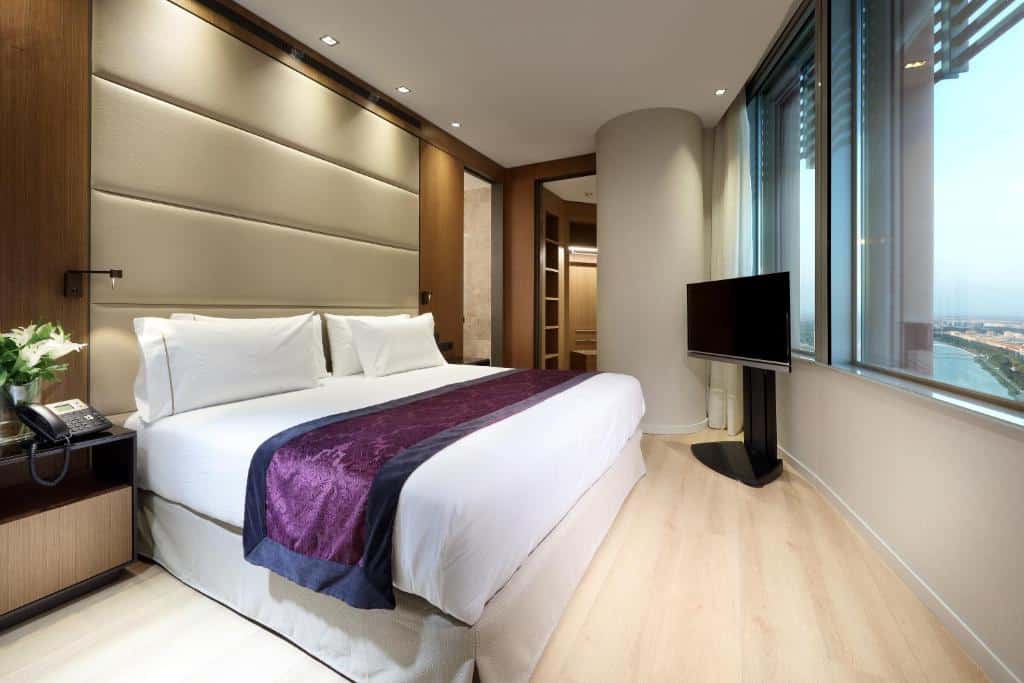 Quarto do Eurostars Torre Sevilla. Uma cama de casal no lado esquerdo e de frente uma janela em toda a parede com a vista pra cidade. Uma televisão no fundo, no canto direito. Foto para ilustrar post sobre Hotéis em Sevilha.