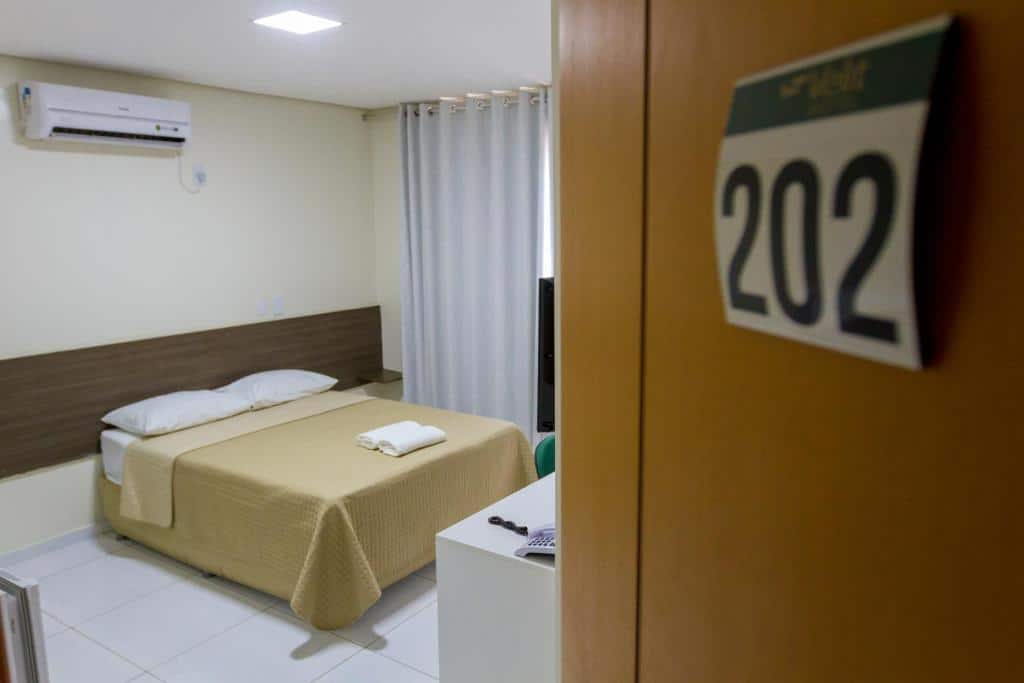 Porta com quarto ao fundo com cama de casal, decoração simples, ar-condicionado e cortinas.