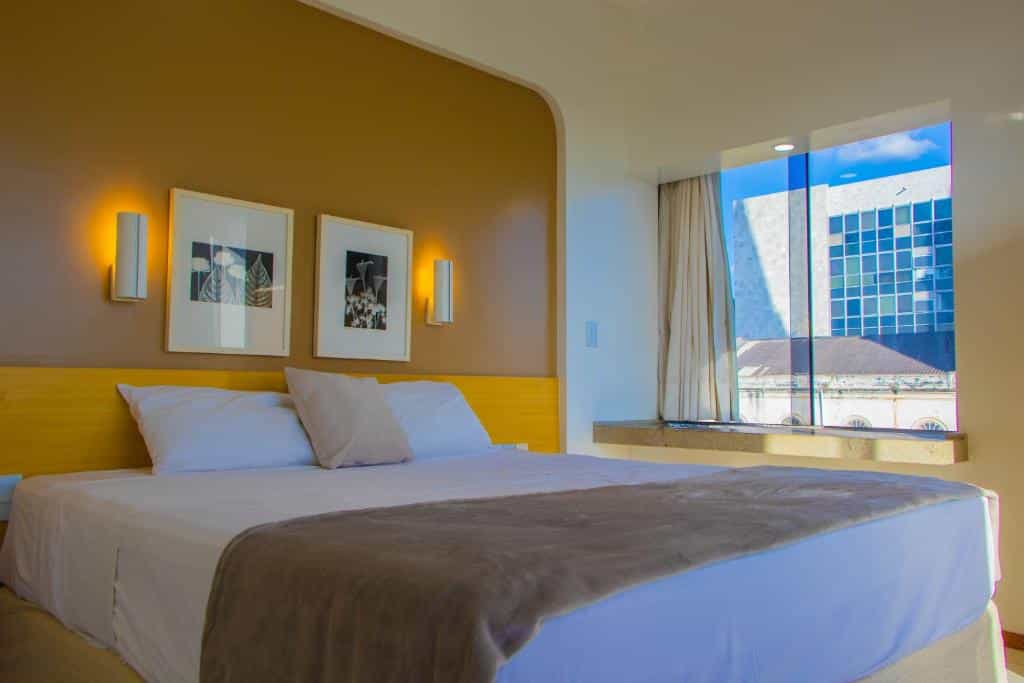 Quarto de hotel com parede amarela, quadros decorativos e janela de vidro com vista para outros edifícios. Imagem para ilustrar o post hotéis em Teresina.