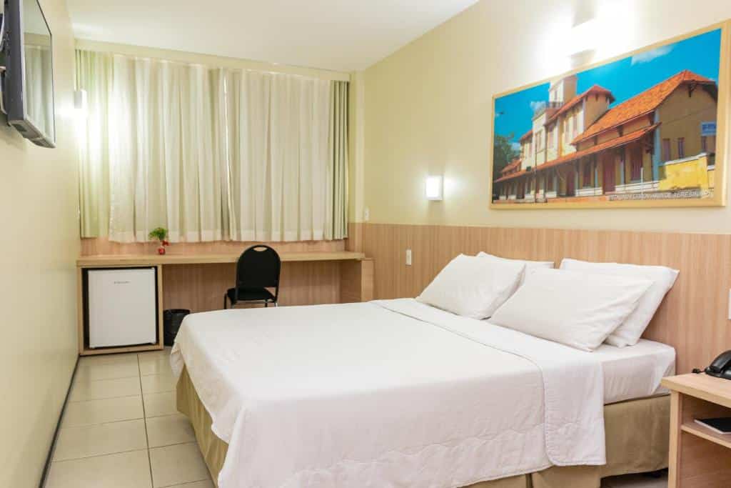 Quarto de hotel com cama de casal, móveis em madeira, quadro decorativo com ponto turístico da cidade, bancada com cadeira e frigobar ao fundo e cortinas. Imagem para ilustrar o post hotéis em Teresina.