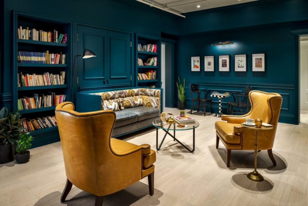Biblioteca do Gladstone House, ambiente com sofás e poltronas, com estantes de livros, tudo decorado em tom de azul escuro, chão de madeira e vasos de flores