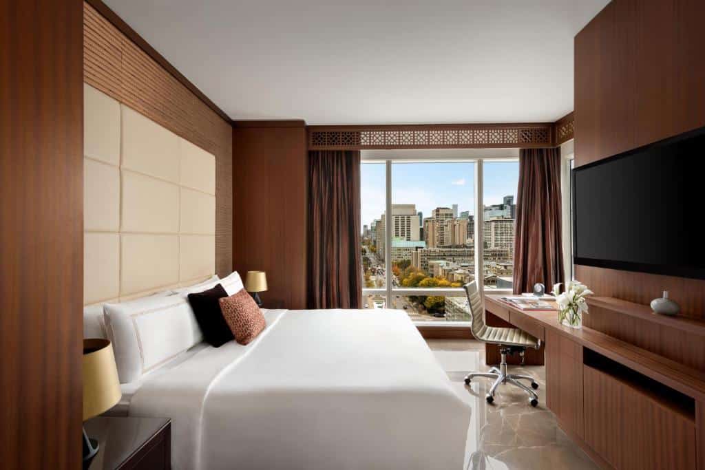 Quarto no Shangri-La com uma janela ampla com vista para a cidade, uma mesa de escritório com uma cadeira, uma cama de casal com travesseiros e almofadas e uma televisão, tudo em madeira e tons de marrorm