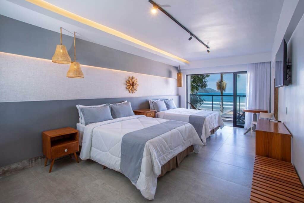 Quarto do Hotel Village Enseada, um dos hotéis em Ubatuba, sendo ele moderno com duas camas de casal, mesas de cabeceira, uma TV na parede e uma varanda com vista para o oceano.