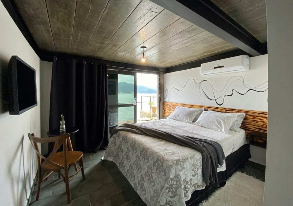 Quarto moderno do 1819 Marine Hotel, um dos hotéis em Ubatuba, com uma cama grande, arte na parede, uma TV, uma cadeira de madeira e uma janela com vista para a montanha.