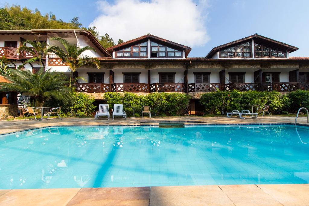 Uma piscina azul clara do Hotelare Hotel Villa Di Capri, um dos hotéis em Ubatuba, em frente a um prédio de dois andares com varandas e espreguiçadeiras em um dia ensolarado.