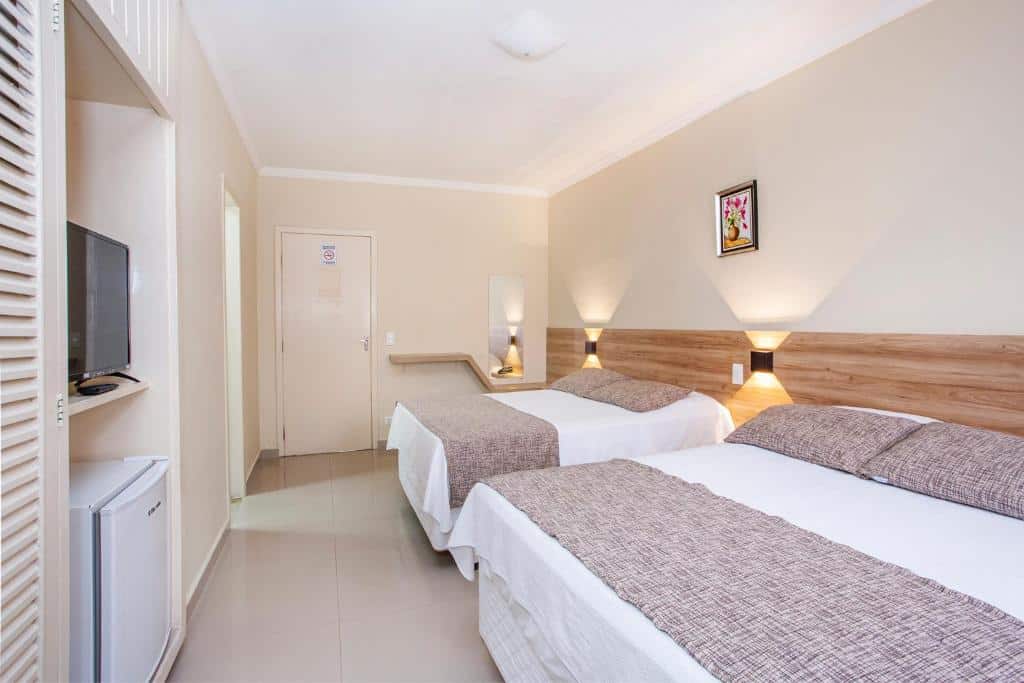 Quarto do Hotel Saveiros, em Ubatuba, com duas camas de casal, uma TV na parede, um frigobar, luminárias e uma foto emoldurada acima das camas.