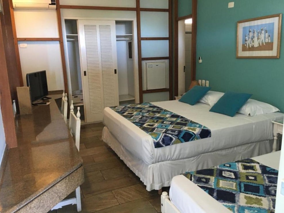 Quarto do Ubatuba Palace Hotel com duas camas, parede com detalhes em azul, escrivaninha com TV, armário aberto e obras de arte emolduradas acima das camas.