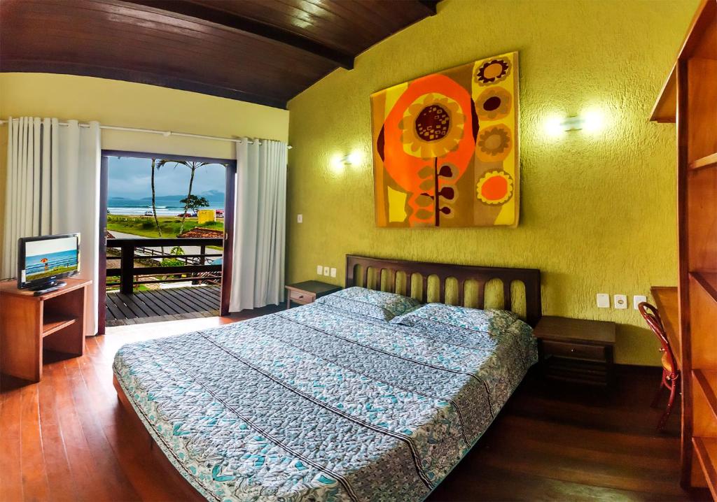Quarto do Hotel Pousada do Sol, um dos hotéis em Ubatuba, com uma cama grande, uma pintura colorida, uma TV em uma mesa e uma varanda com vista para o oceano e palmeiras.
