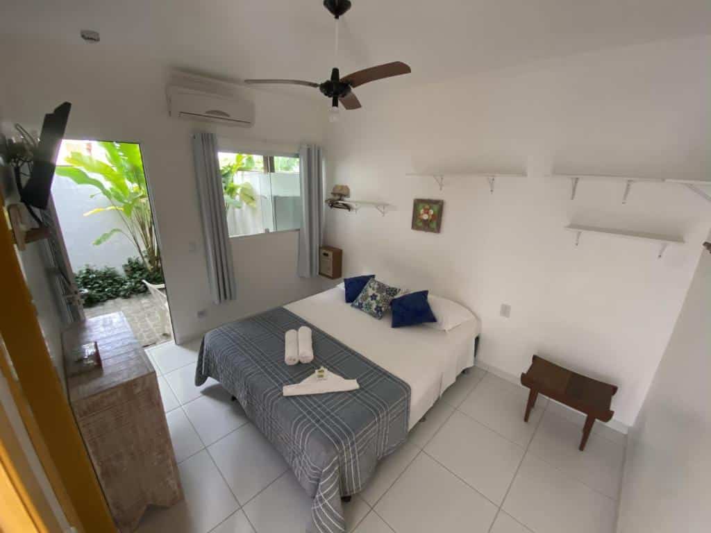 Quarto arrumado do Hotel e Pousada Pouso54, um dos hotéis em Ubatuba, com uma cama de casal, dois travesseiros azuis, um ventilador de teto e uma porta aberta que dá para uma pequena área externa.