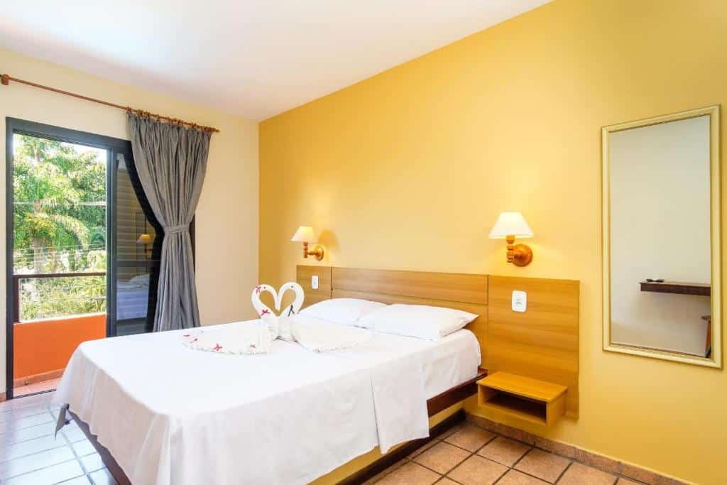Quarto com cama de casal e toalhas em formato de cisne no Hotel Ponta das Toninhas, em Ubatuba. Há uma varanda com vista para árvores verdes