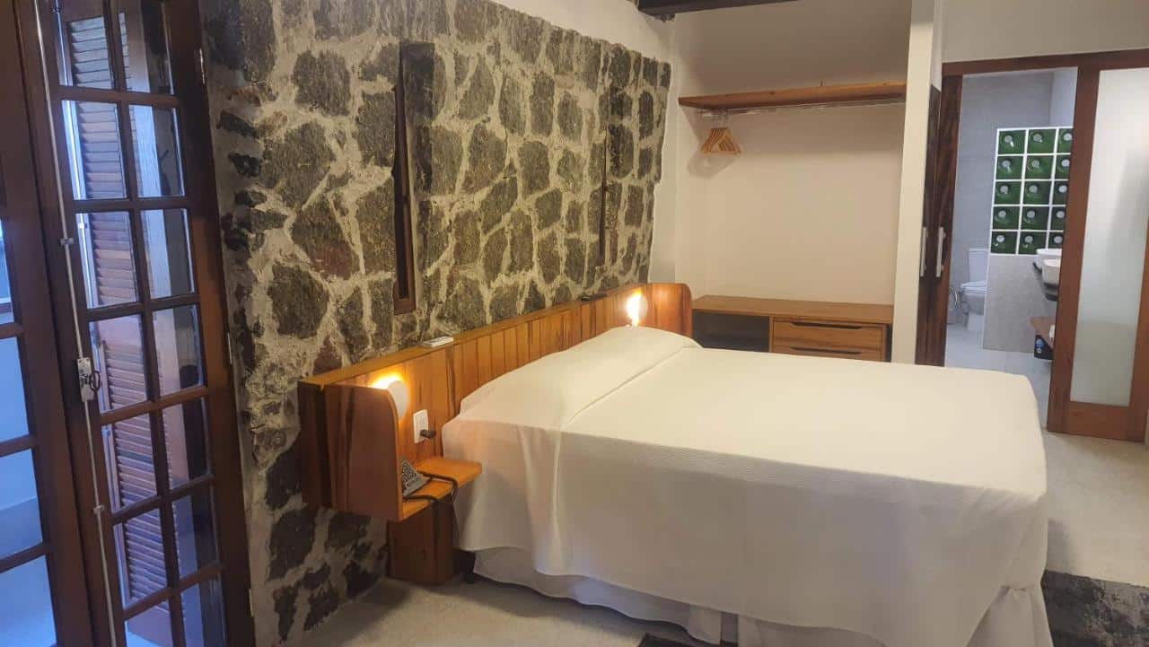 Cama bem arrumada do Hotel Coquille, um dos hotéis em Ubatuba, com cabeceira de madeira contra uma parede de pedra, ao lado de prateleiras abertas e uma porta de vidro.