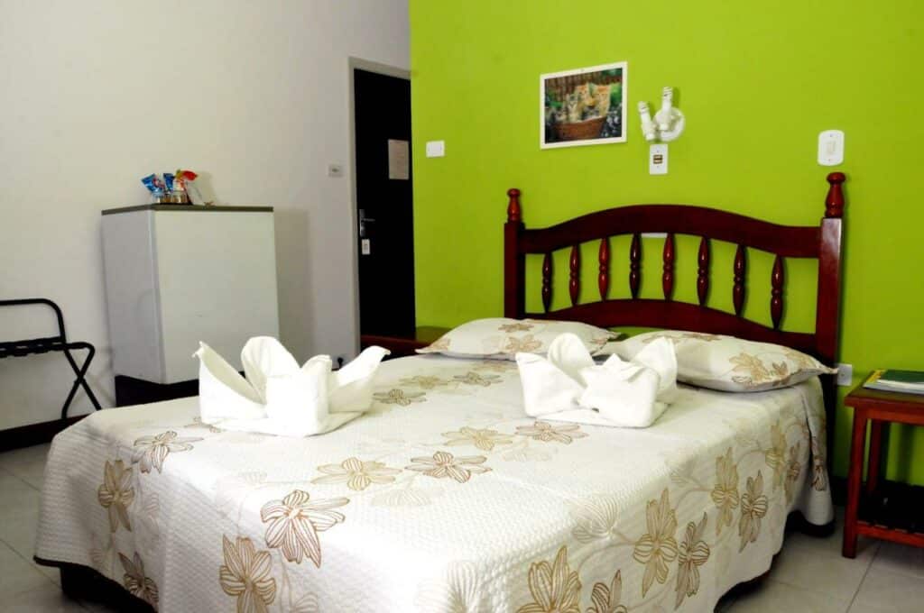Quarto do Hotel São Nicolau, um dos hotéis em Ubatuba, sendo ele arrumado com uma cama de casal, roupa de cama floral, cisnes de toalha, um pequeno frigobar e uma parede verde brilhante.