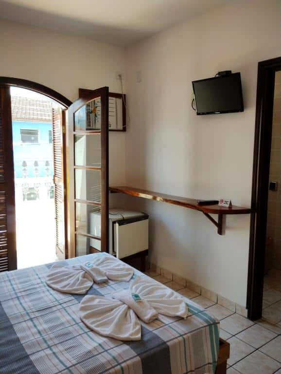 Um pequeno quarto do Hotel Xaréu, um dos hotéis em Ubatuba, com uma cama, TV na parede, frigobar e toalhas dobradas, com uma porta aberta para uma varanda.