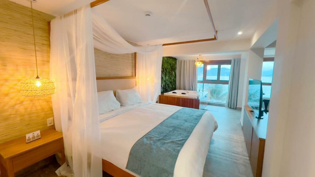 Quarto do Catalina Hotel, em Ubatuba, com cama de dossel, mesa de cabeceira, banheira de hidromassagem, janelas grandes e vista para o oceano.