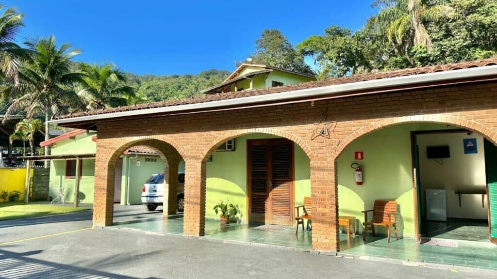 Frente do Hotel Bem Te Vi, um dos hotéis em Ubatuba, com arcos de tijolos, paredes verdes, assentos ao ar livre e um carro estacionado, cercado por palmeiras e vegetação.