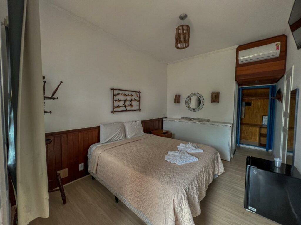 Um quarto simples do Hotel Porto do Eixo, em Ubatuba, com uma cama de casal, roupa de cama bege, arte na parede, um espelho e um ar-condicionado na parede.