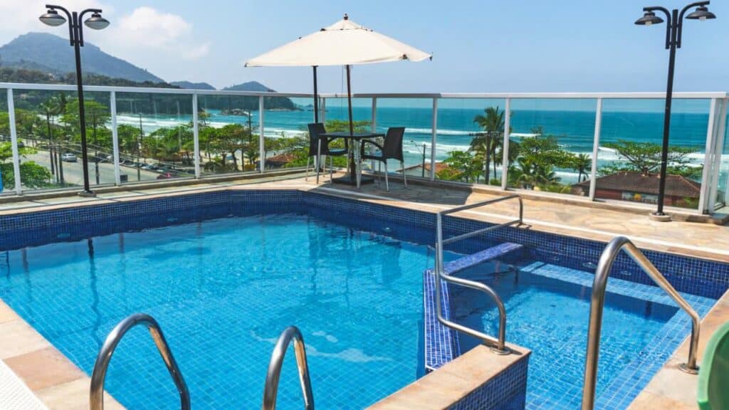 Piscina na cobertura do UPG Hotel, em Ubatuba, com degraus, uma mesa e um guarda-sol tem vista para a praia e o oceano sob um céu claro.