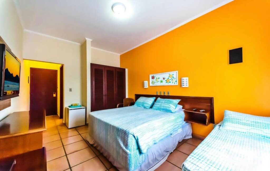 Quarto do Hotel Villa Di Rimini, um dos hotéis em Ubatuba, com uma parede com detalhes em laranja, uma cama de casal, uma cama de solteiro, uma TV, uma escrivaninha e um pequeno frigobar.