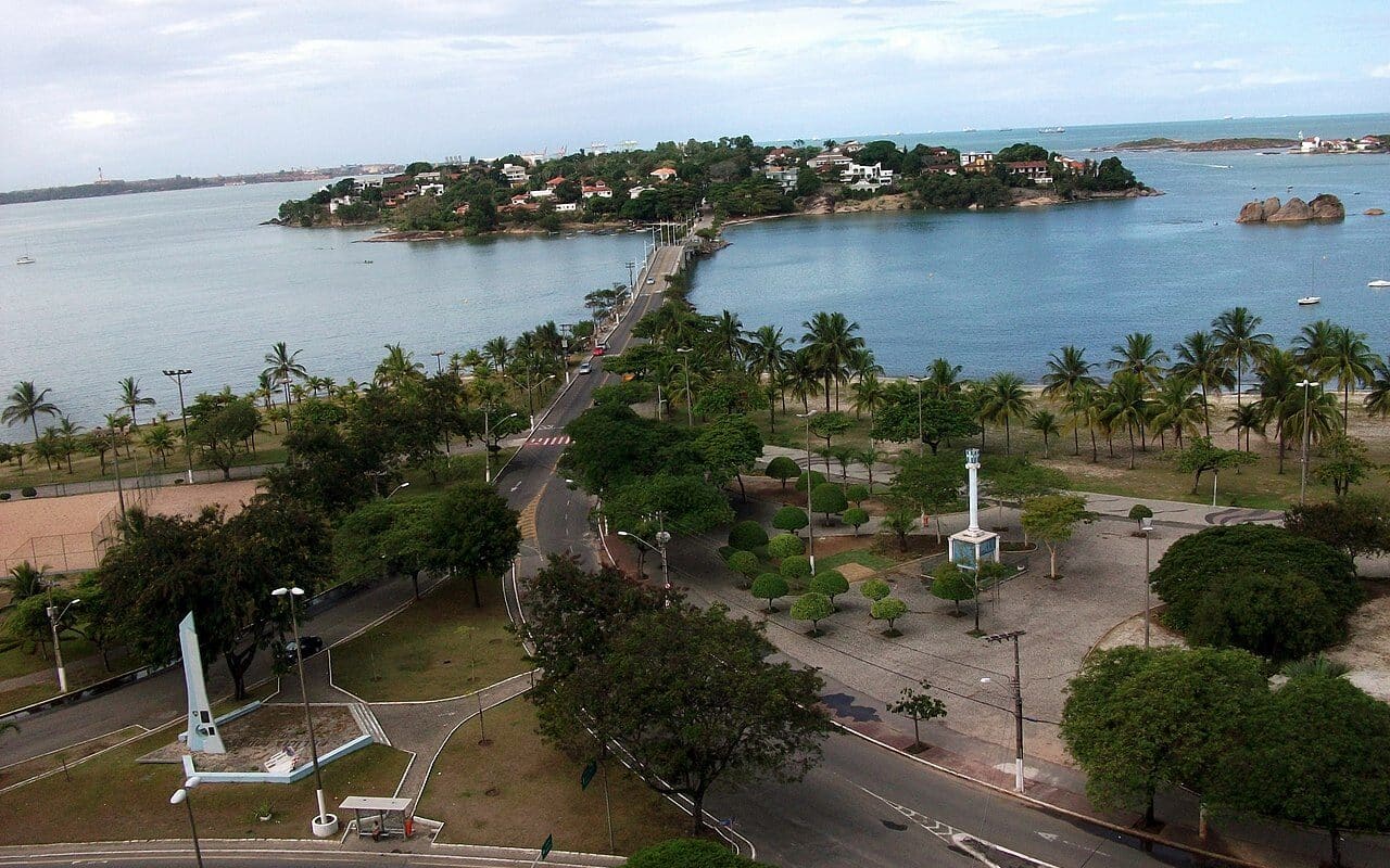 Vista de Vitória com muitas árvores e a ponte levando até a ilha, com várias casas e árvores. É possível ver o mar, rochas e alguns barcos no mar.