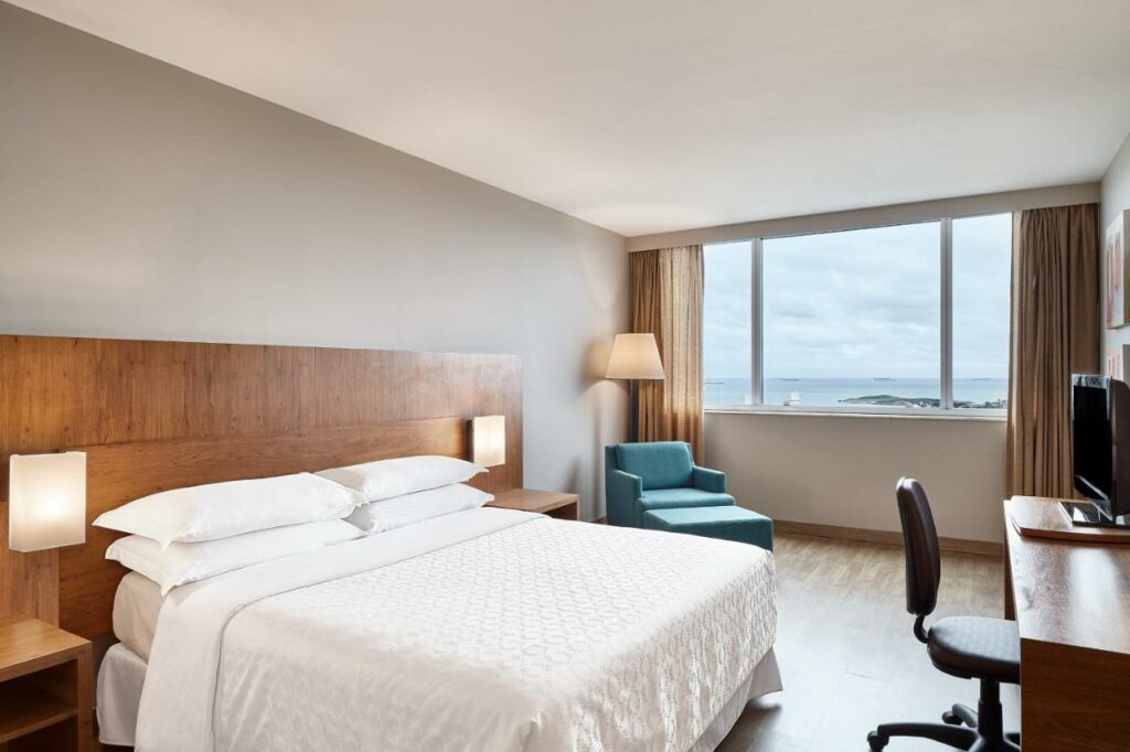 Quarto no Sheraton Vitoria com uma cama de casal, mesa com cadeira, televisão, poltrona e uma janela com cortinas e vista da praia, para representar hotéis em Vitória.