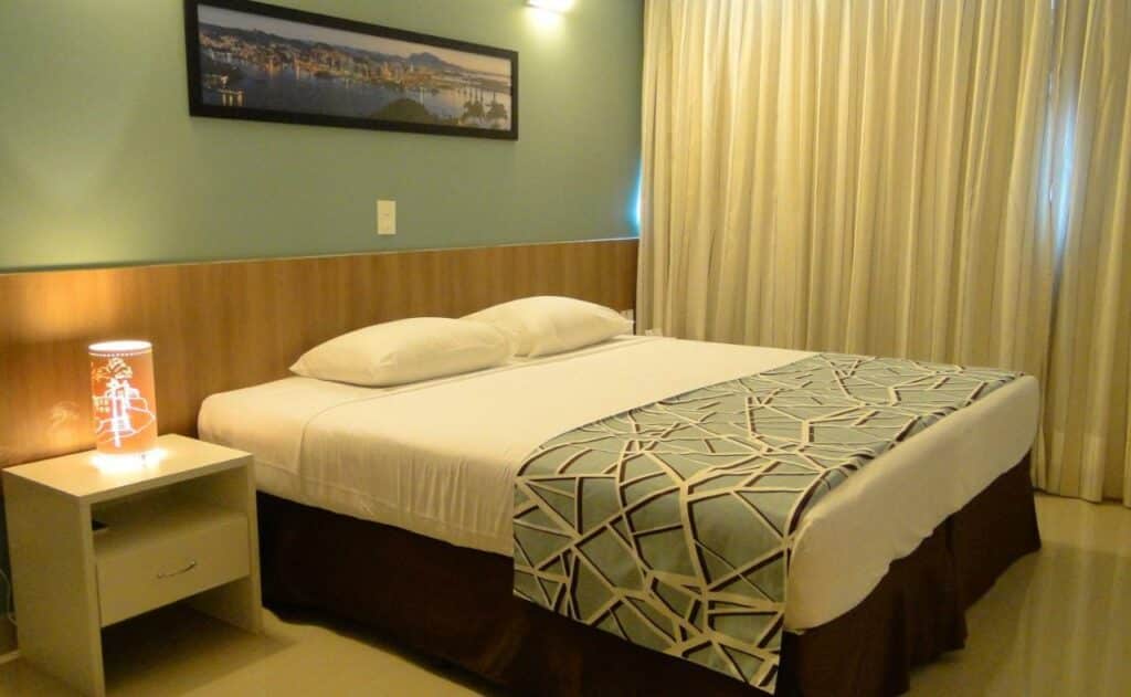 Quarto no Alameda Vitória Hotel com uma cama de casal, mesinha com abajur e janela com cortina, para representar hotéis em Vitória.
